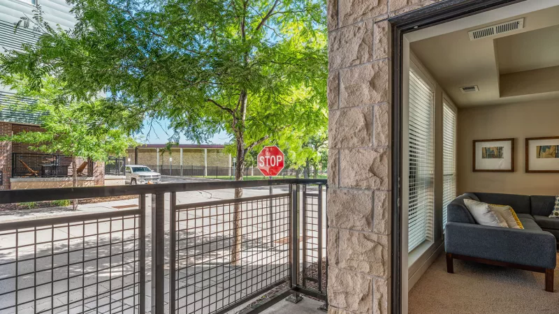 Balcony with metal railing, overlooking a tree-lined street, and an interior view of a living room with a gray sofa.