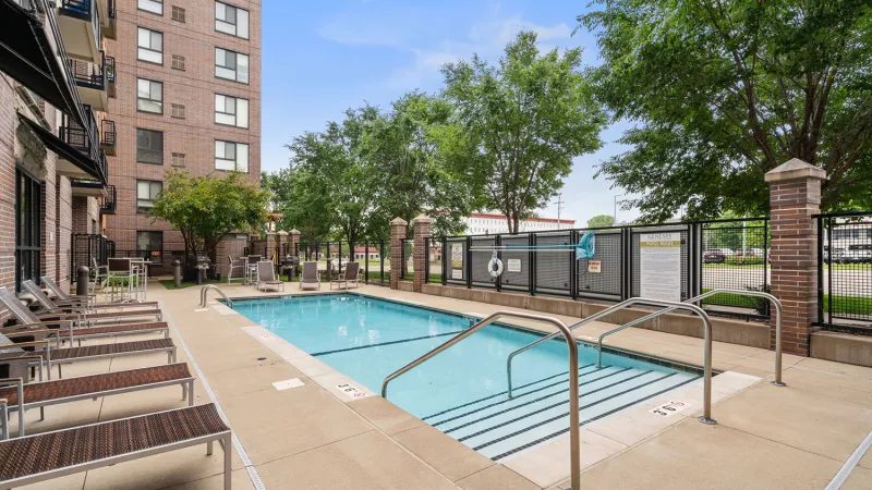 Outdoor swimming pool with lounge chairs next to a modern brick apartment building and green trees under a blue sky.
