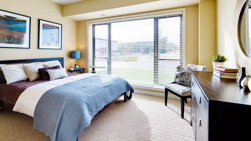 Bright bedroom with light yellow walls, a bed with a blue blanket, and a large window overlooking green space.