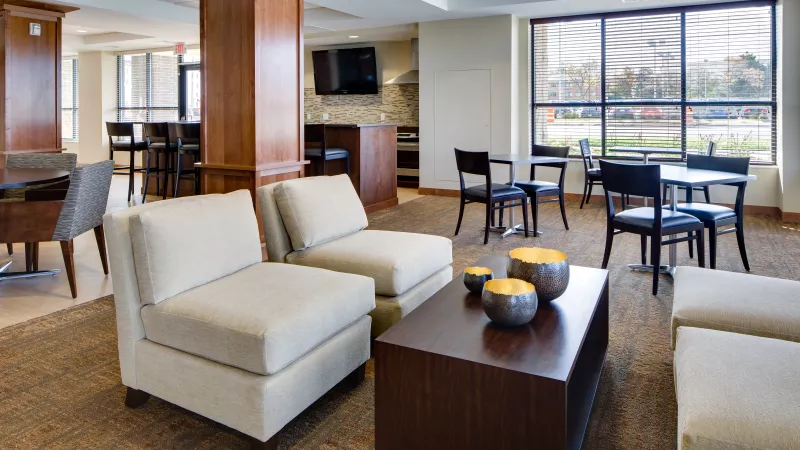 Spacious lounge with beige armchairs, a coffee table, and small dark tables near large windows.