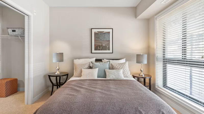 A neutral-toned bedroom with a queen bed, two nightstands with lamps, a window with blinds, and an open closet.