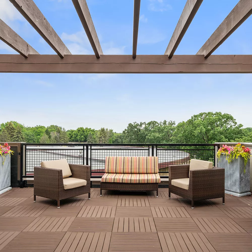 Outdoor patio with a brown wicker sofa, two armchairs, and flowering planters under a wooden pergola, overlooking a green landscape.