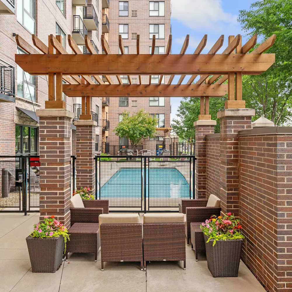 Outdoor patio with brown wicker furniture, wooden pergola, and potted flowers. A swimming pool and apartment buildings in background.
