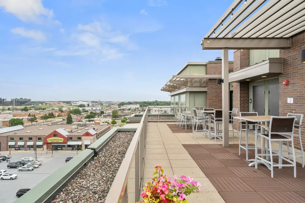 Rooftop patio with high-top tables, chairs, and potted flowers overlooking a city skyline under a blue sky.