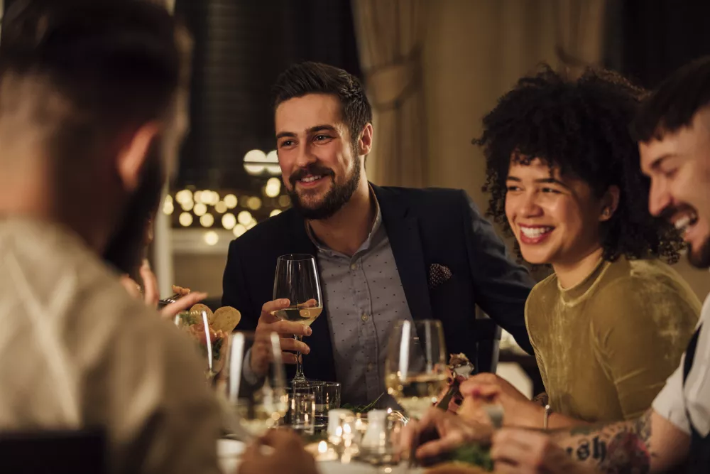 Four happy friends, including a man and woman holding wine glasses, share a lively dinner at a dimly lit restaurant.